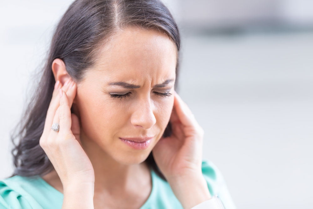 Young brunette woman with her hands to her ears, potentially struggling with ear pain, ear infection, or earwax impaction.