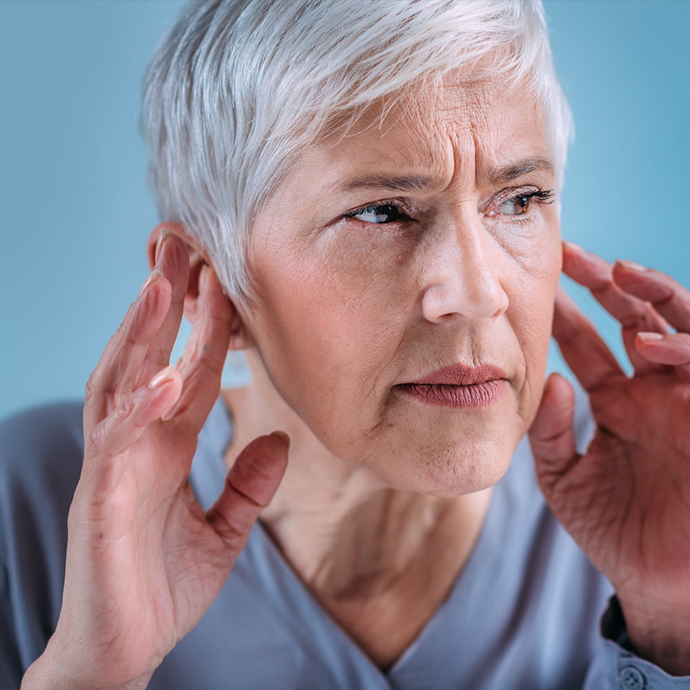 Elderly woman touching her ears with the tips of her fingers, visibly struggling to hear.