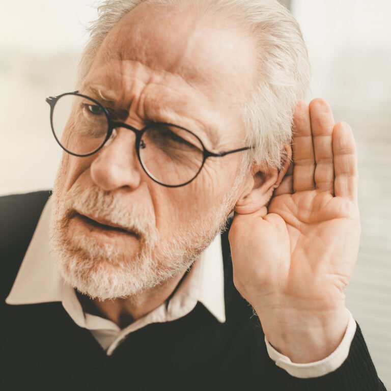 Senior man with white hair and glasses holding his hand to his left ear, visibly struggling to hear.
