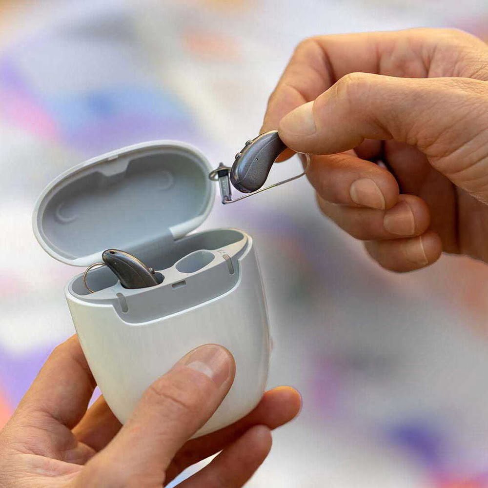 Closeup of hands holding a hearing aids charging case, inserting hearing aid into slot.