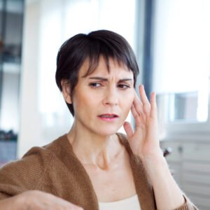 Middle-aged woman with her left hand raised to her ear, visibly in discomfort, potentially suffering from tinnitus or hearing loss.